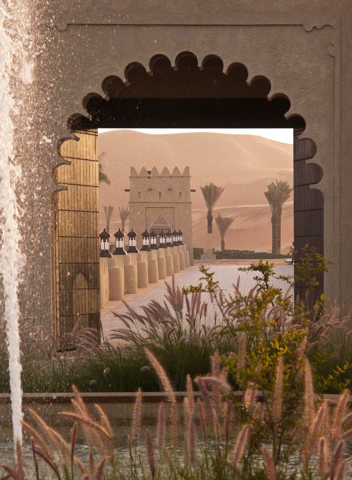 Arched stone doorway of a desert resort frames a desert landscape. A pathway lined with lanterns leads to a distant fort, with sand dunes and palm trees beyond.