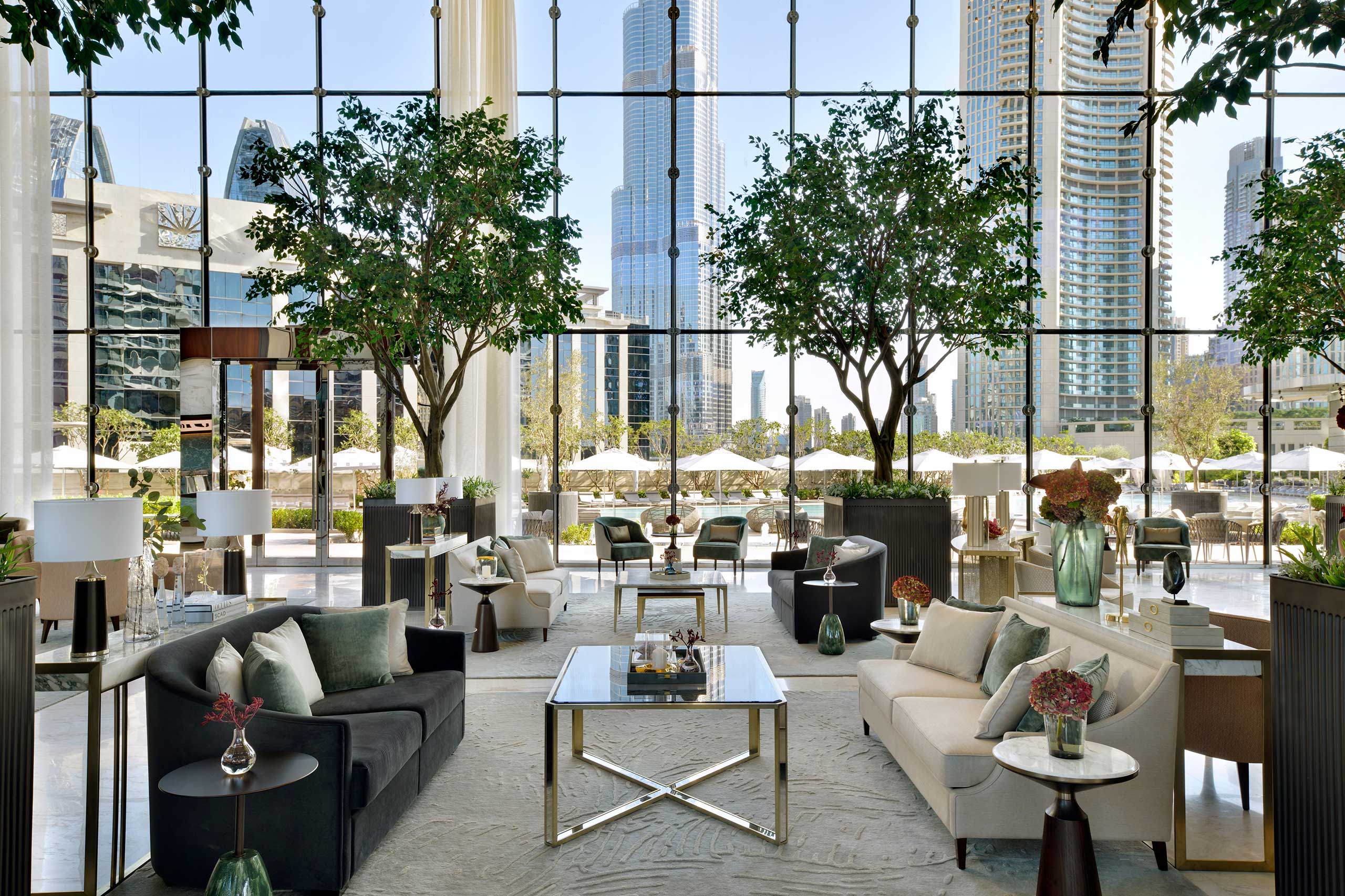 Luxurious hotel lobby with modern sofas, tables, and lush indoor trees. Floor-to-ceiling windows reveal a cityscape with tall skyscrapers. Bright and elegant atmosphere.