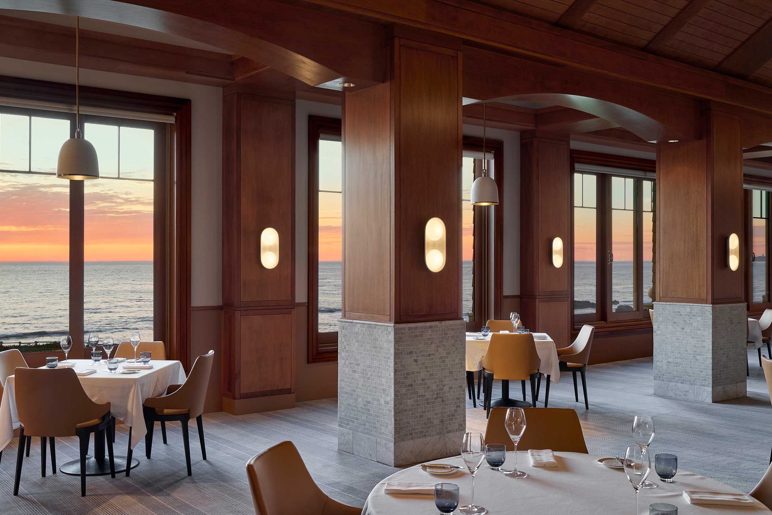 Elegant restaurant interior with ocean-view windows at sunset. Warm wood tones, white tablecloths, and modern lighting create a serene and inviting atmosphere.
