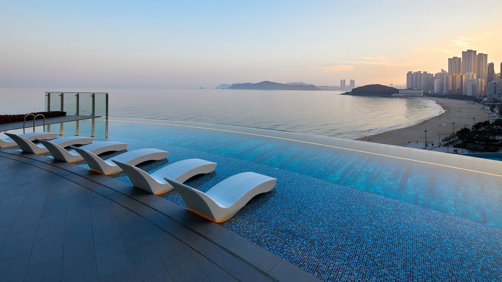 Infinity pool of a resort with sleek white loungers overlooks a calm ocean, distant city skyline, and a serene beach at sunset, creating a tranquil, luxurious vibe.