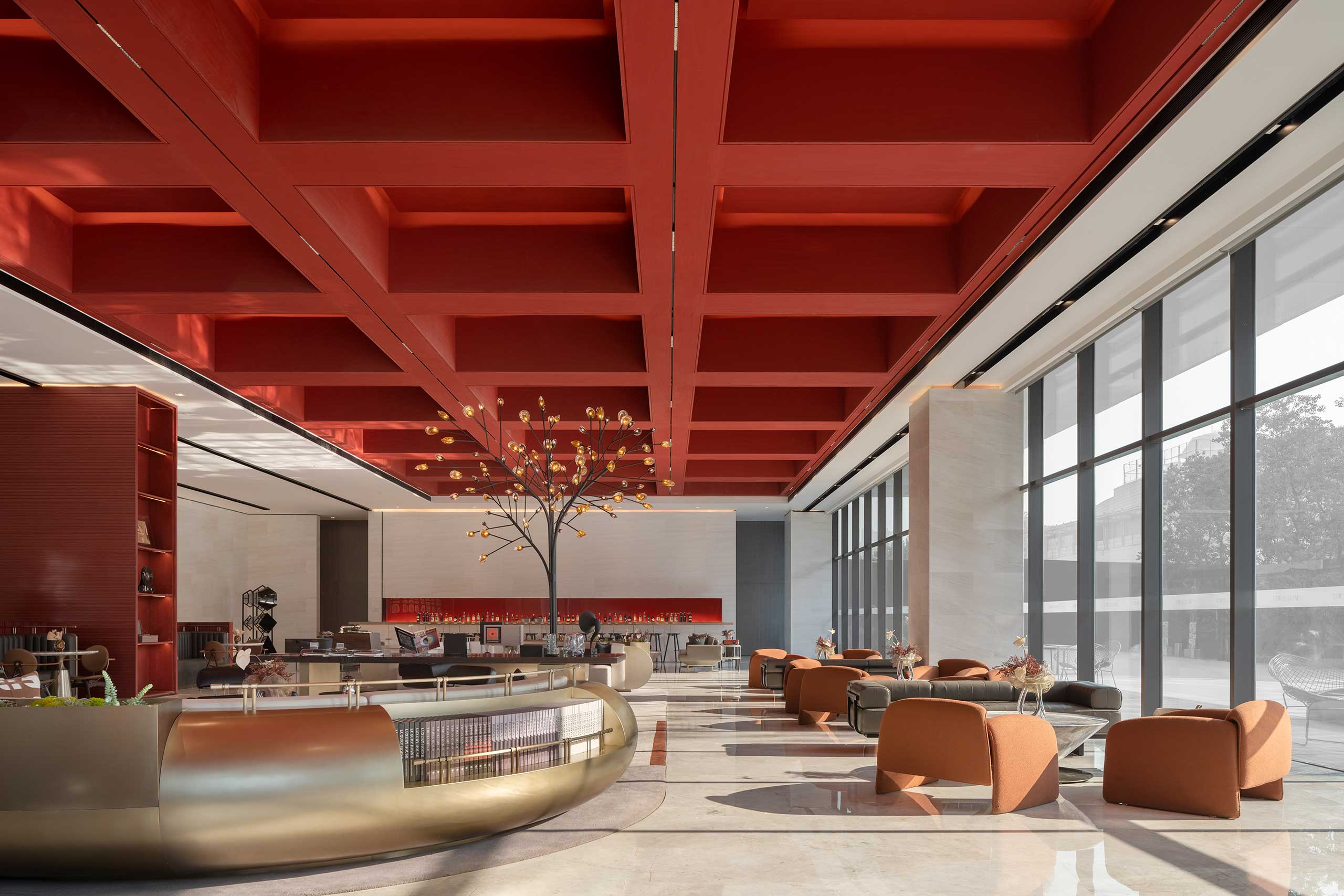 Spacious modern office lobby with high red ceiling, large windows, and chic seating areas. A decorative tree sculpture adds elegance and warmth.