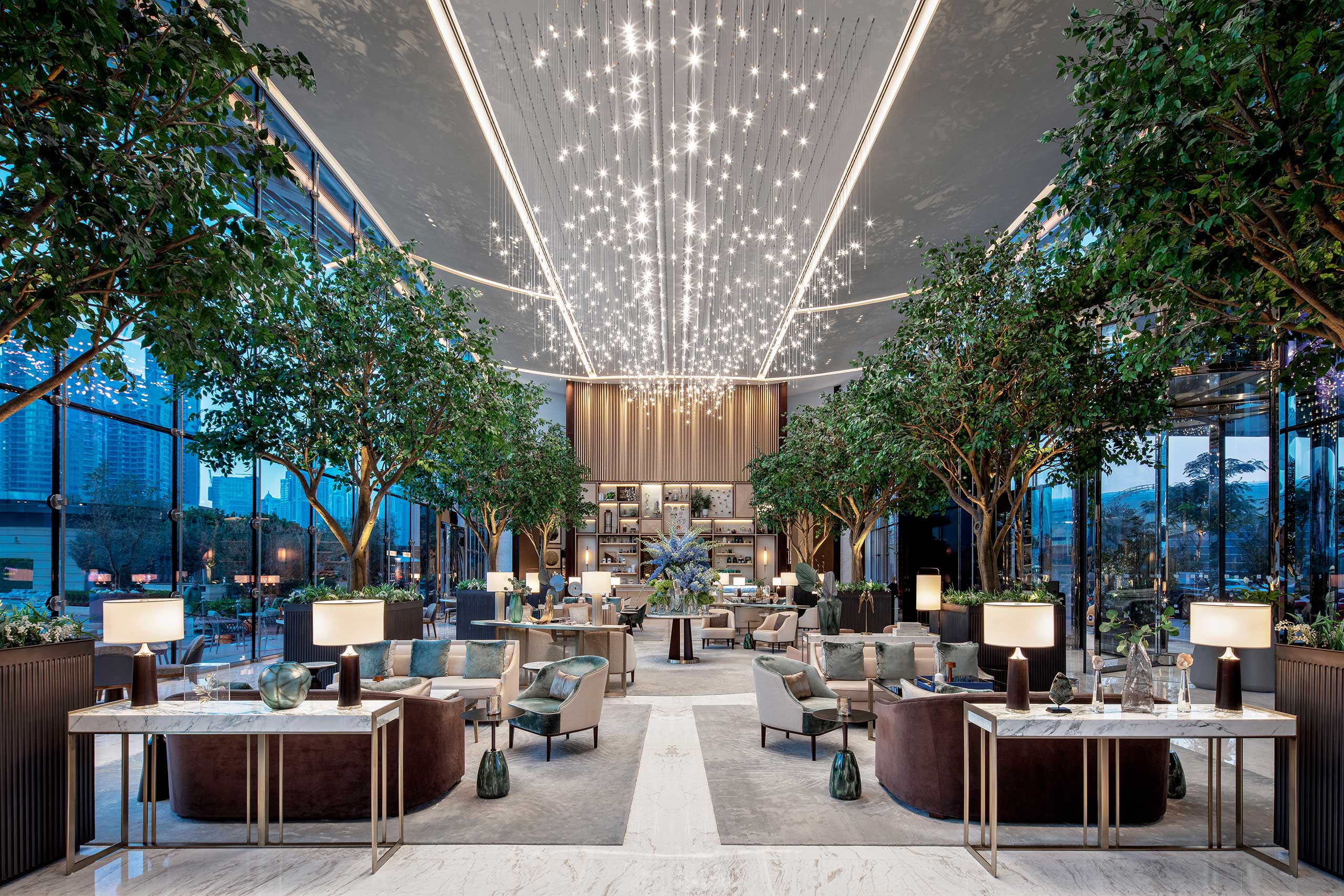 Luxurious hotel lobby with high ceiling adorned with star-like lights, flanked by tall indoor trees. Elegant seating and modern decor create a serene ambiance.