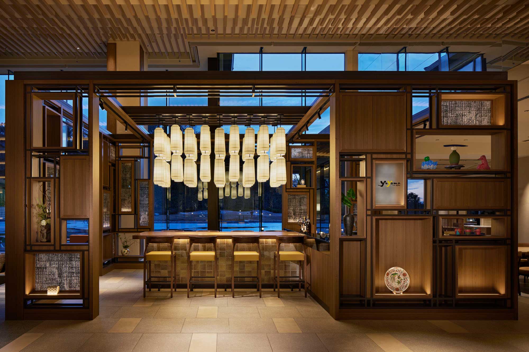 Warmly lit modern hotel bar with wooden accents, hanging lanterns, and five stools. A large window offers a view of trees against a dusky sky. Cozy and inviting ambiance.