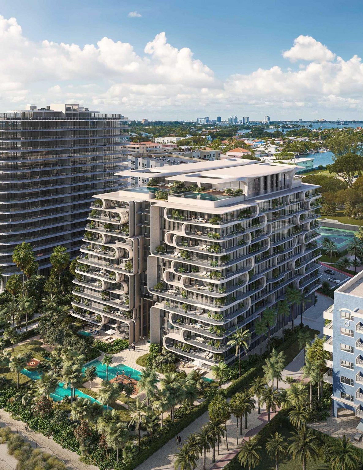 Aerial view of modern luxury condos with distinctive curved balconies. Surrounded by palm trees and pools, with a calm waterway and city skyline in the distance.