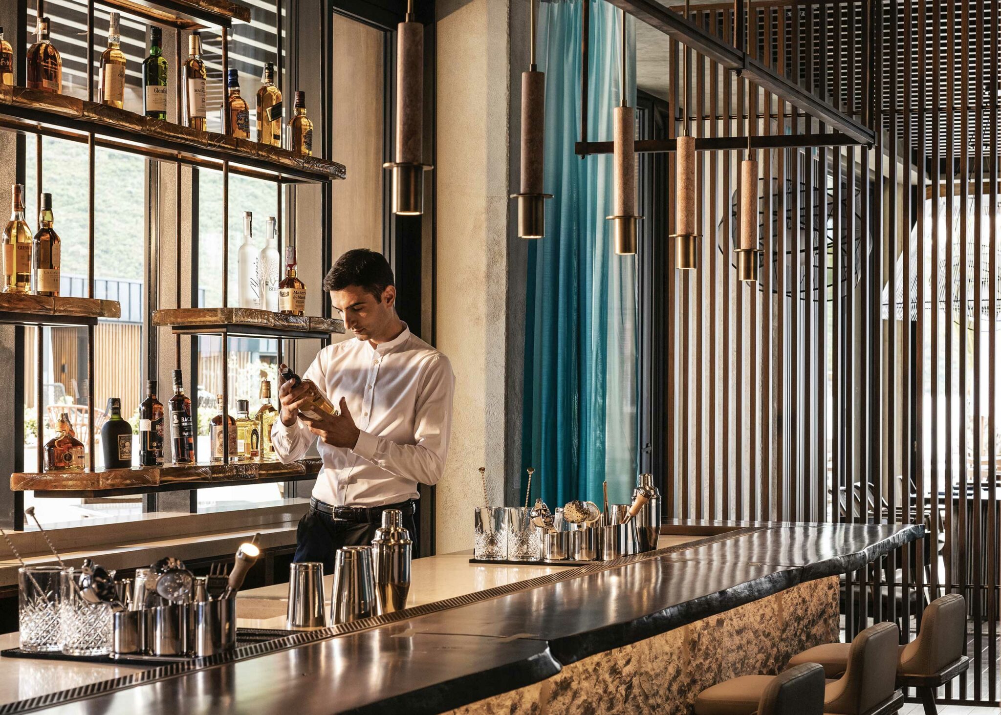 A bartender in a white shirt examines a bottle behind a sleek, modern bar with various bottles on shelves. The scene is warmly lit and stylish.