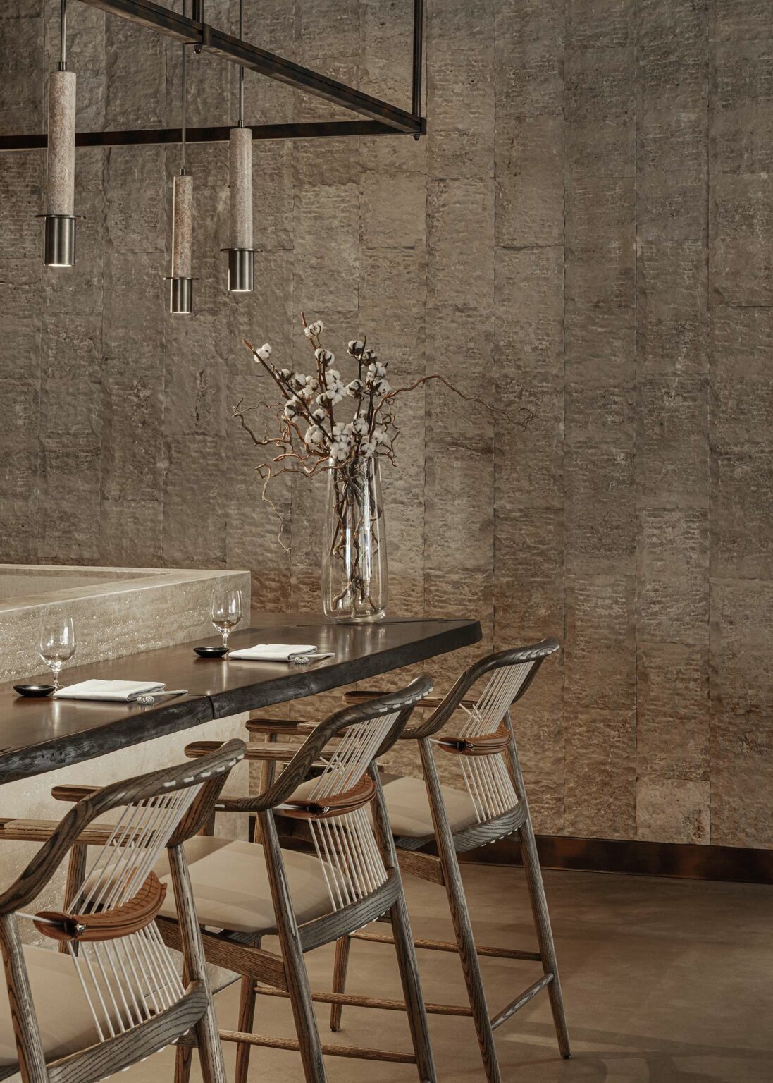 Sophisticated dining area with woven chairs and a sleek wooden table set with glasses. Elegant pendant lights and a vase of cotton branches add charm.