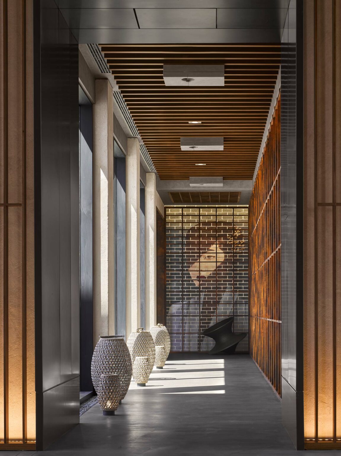Modern hallway with large windows casting shadows. Three textured vases line the floor. A brick wall features a partial face portrait, enhancing the elegant, serene ambiance.