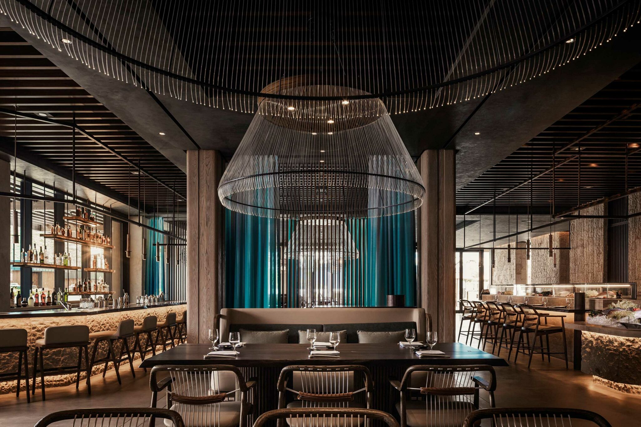 Elegant restaurant interior with a modern design, featuring large, intricate chandeliers, teal curtains, a fully stocked bar, and neatly set tables and chairs.