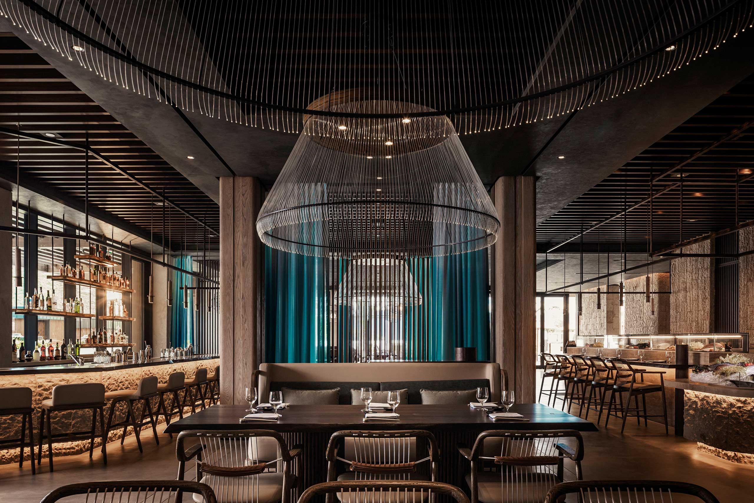 Elegant restaurant interior with a modern design, featuring large, intricate chandeliers, teal curtains, a fully stocked bar, and neatly set tables and chairs.