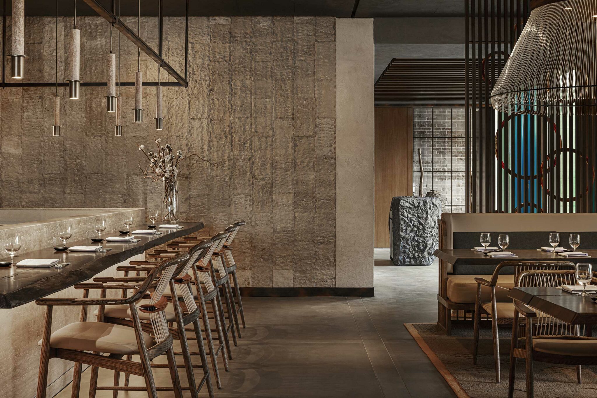 Elegant restaurant interior with a modern, minimalist design. Features wooden chairs, stone-textured walls, pendant lights, and set dining tables.