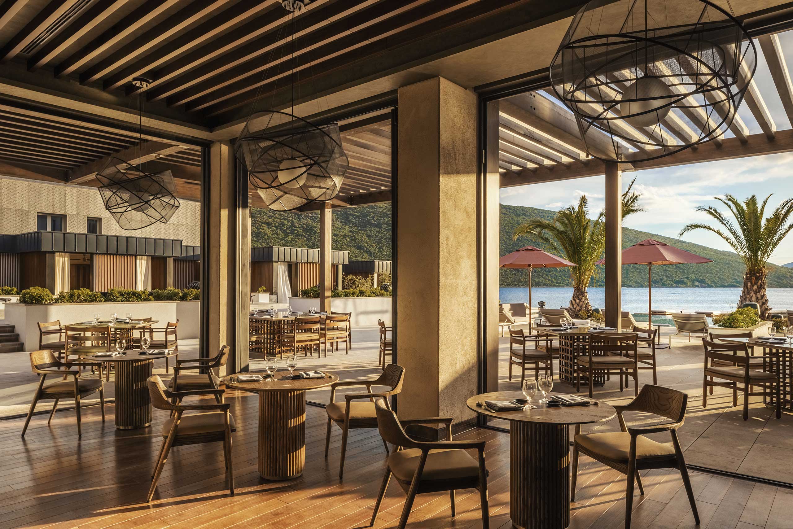 Elegant open-air restaurant with wooden tables and chairs, under geometric chandeliers. Large windows reveal mountains, palm trees, and umbrellas by the sea.