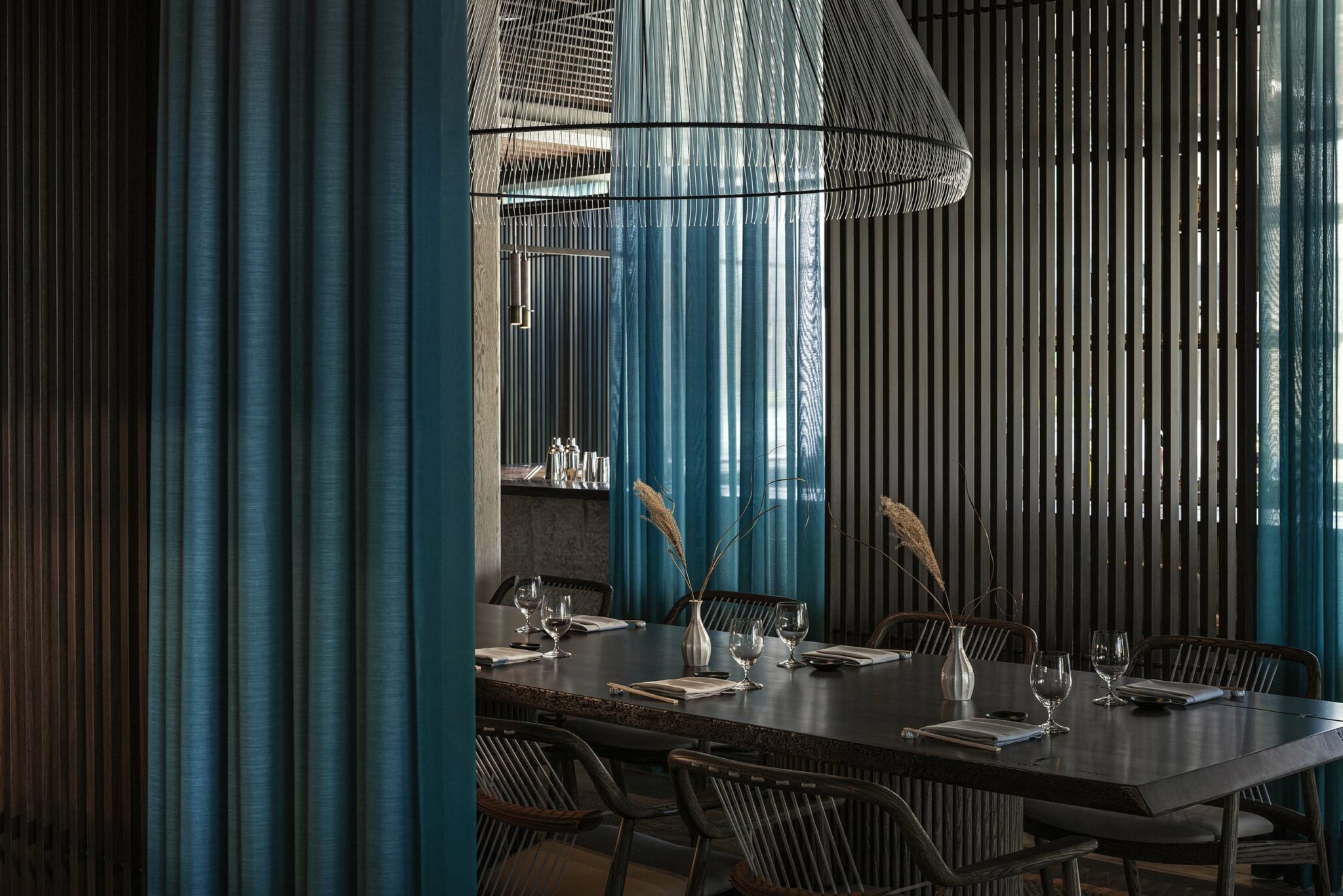 Sophisticated dining room with a dark wood table set for eight. Elegant blue curtains and sleek vertical slats create a modern, serene ambiance.