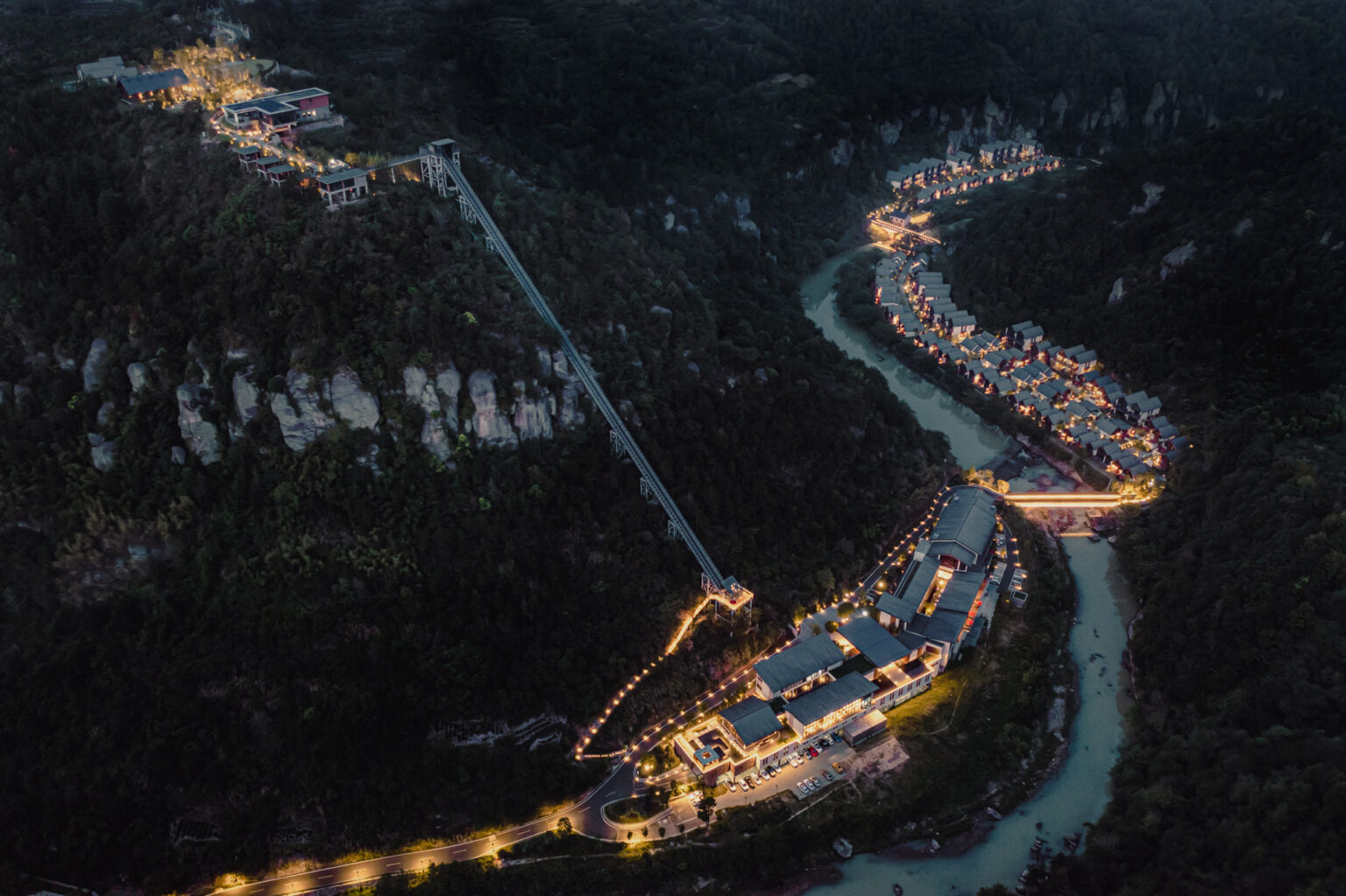 A nighttime aerial view of a mountain village, featuring a winding river and illuminated buildings. A lit cable car connects the ridge to the valley.