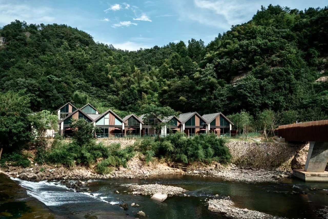 Modern cabins with A-frame roofs are nestled against a lush green hillside, overlooking a gentle river. The scene conveys tranquility and natural beauty.