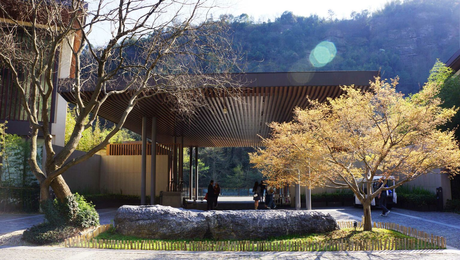 Modern hotel pavilion with a slatted roof set against a hillside. Leafless trees and golden foliage add contrast. People gathered under dappled sunlight.