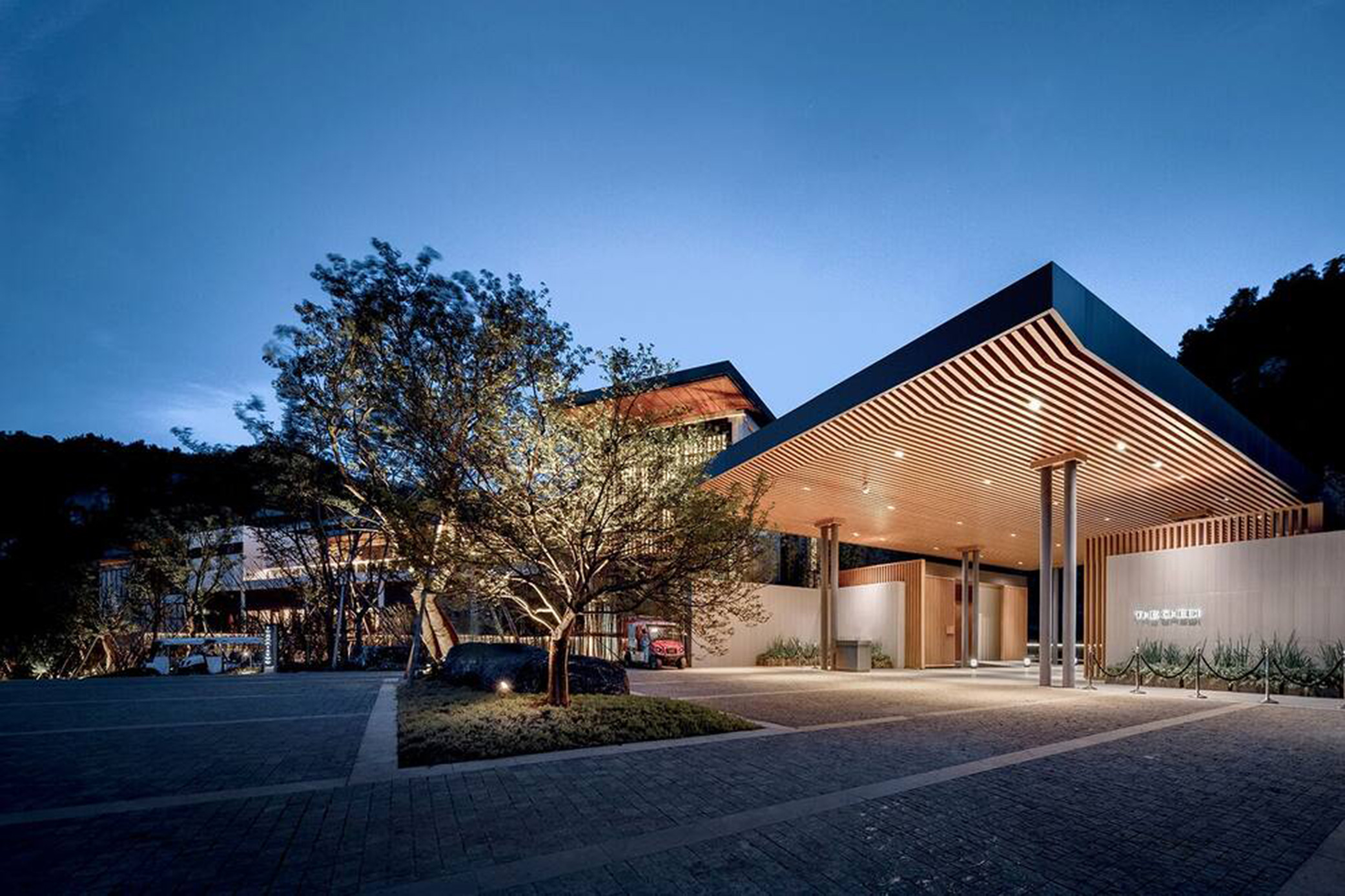 Modern hotel building with a sleek wooden overhang and illuminated entrance at dusk. A tree is artistically lit, creating a serene and inviting atmosphere.