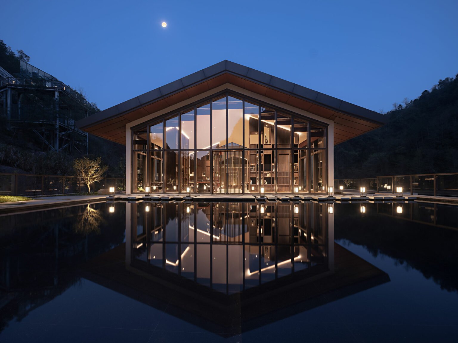 Modern glass pavilion at dusk, softly illuminated, reflects in a serene water pool. Surrounded by trees, it conveys elegance and tranquility.