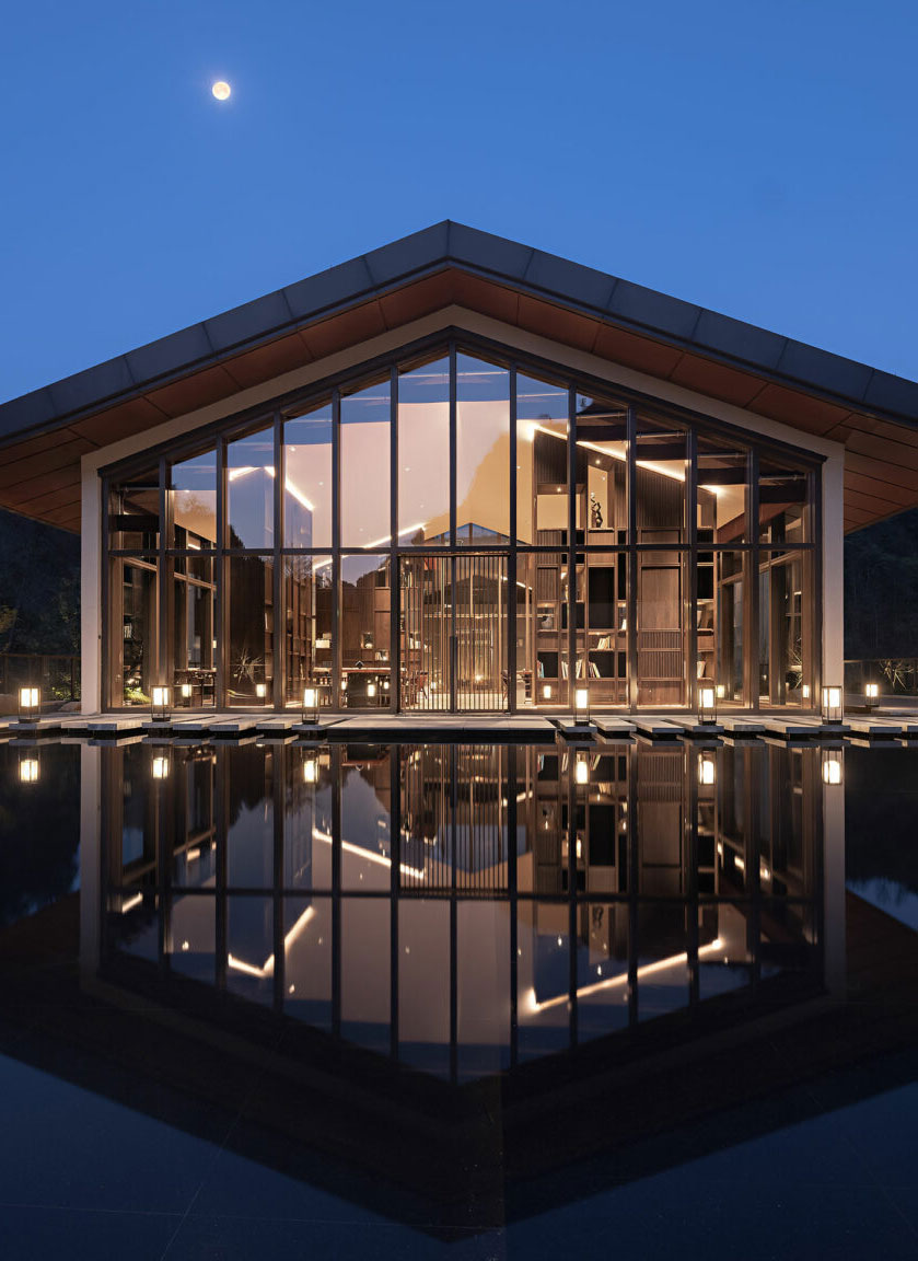 Modern glass pavilion at dusk, softly illuminated, reflects in a serene water pool. Surrounded by trees, it conveys elegance and tranquility.