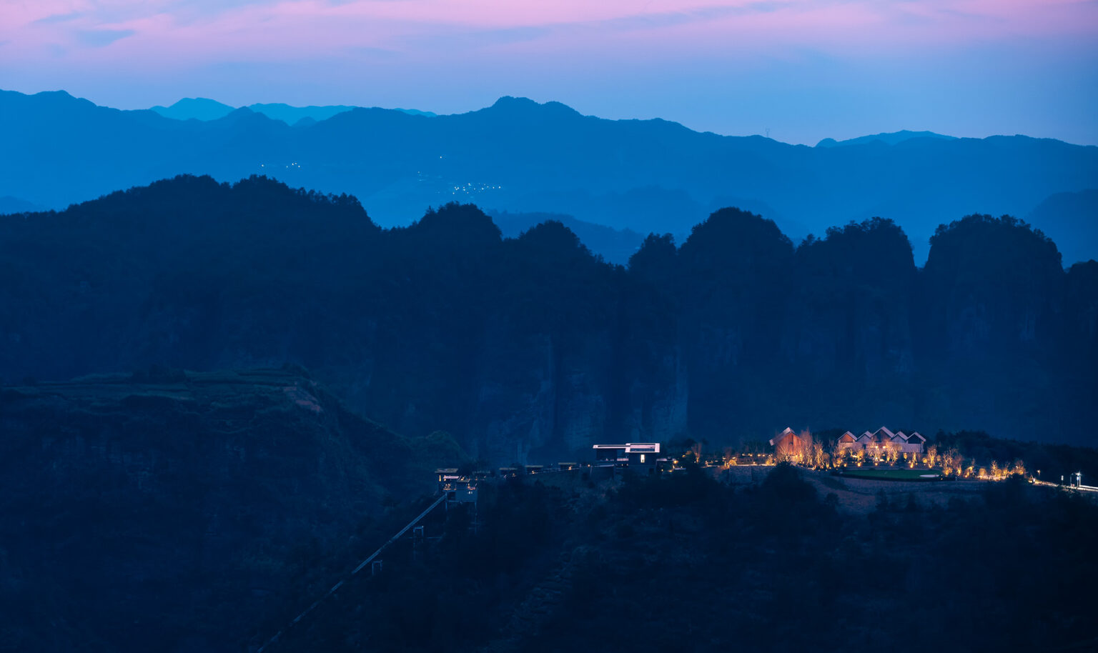 Mountainous landscape at dusk with silhouetted peaks, a softly lit hotel, and glowing trees. A tranquil, serene atmosphere under a pink-tinged sky.