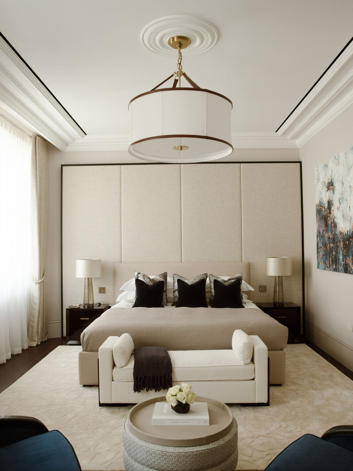Elegant bedroom with neutral tones. Features a large bed with black pillows, flanked by bedside lamps. A plush bench and round table add a cozy touch.