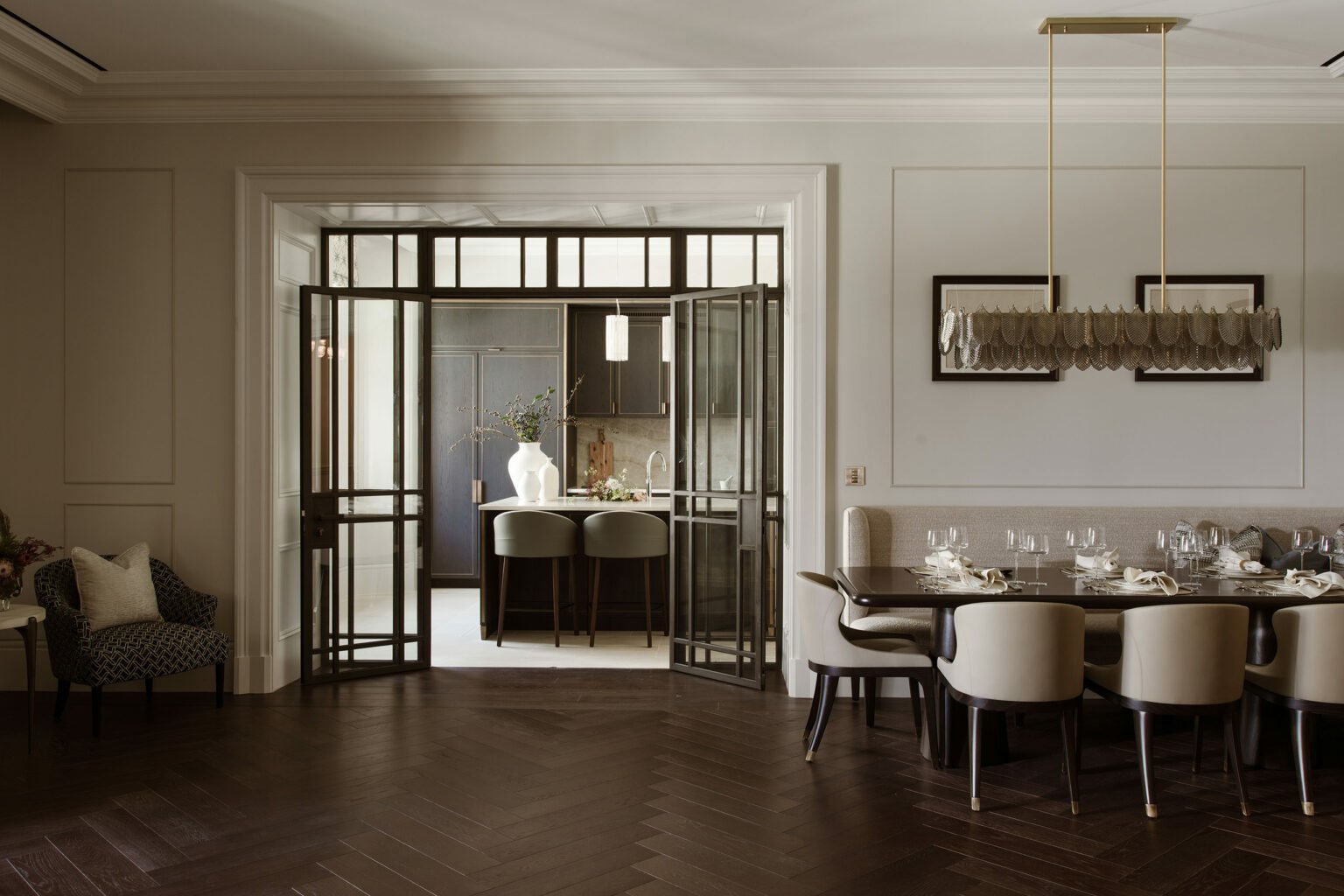 A sophisticated dining room with herringbone wood flooring, a dark wood table set for dinner, and cream chairs. An elegant modern chandelier hangs above, and black framed glass doors lead to a kitchen visible in the background.