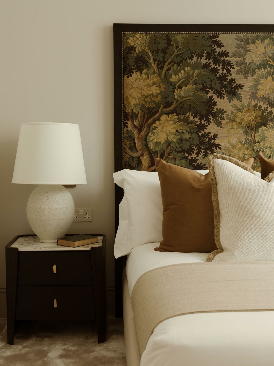 Elegant bedroom scene with a tapestry headboard depicting trees, a neatly made bed with brown and white pillows, and a bedside table with a lamp and book. Calm, cozy atmosphere.