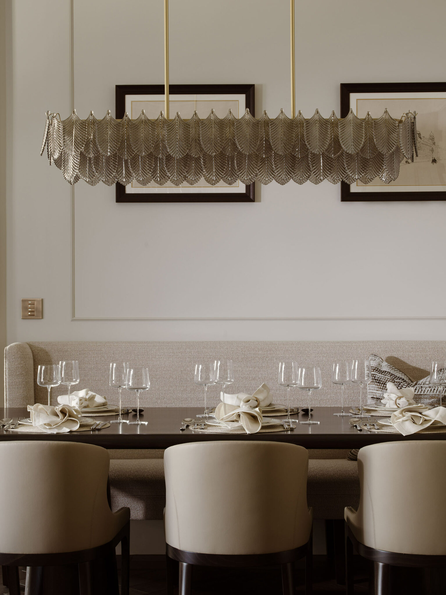 Elegant dining room with a metallic chandelier, six upholstered chairs, set table with glasses and napkins. Neutral tones and framed artwork on walls.