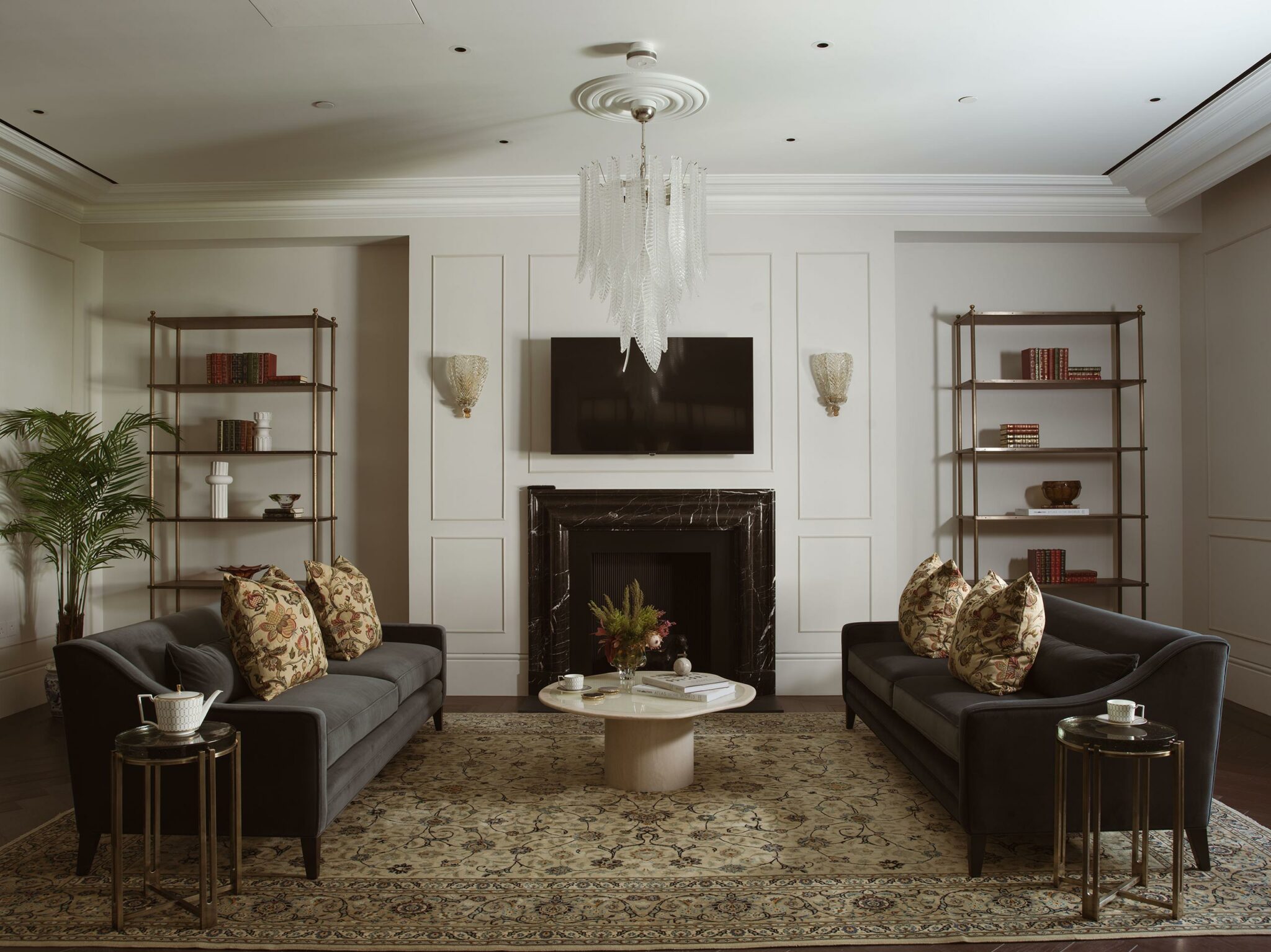 Elegant living room with two dark sofas facing each other, floral cushions, a central marble coffee table, ornate chandelier, built-in bookshelves, and fireplace.
