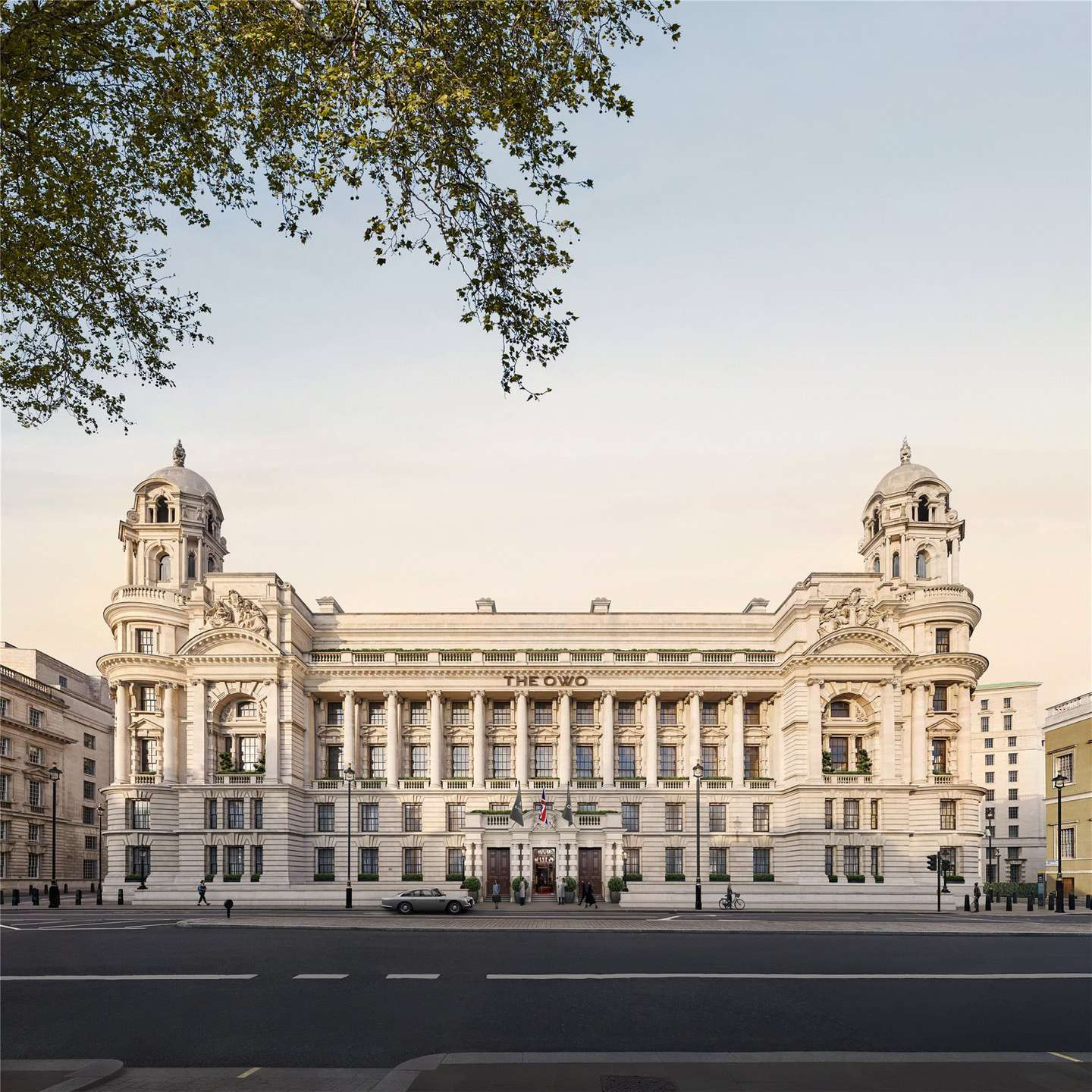 The OWO Residences by Raffles exteriors. Majestic neoclassical building with grand columns, ornate facades, and two towers; the setting is serene with a pale sky and sparse traffic.