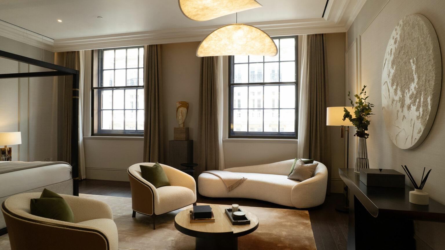 Elegant living room with neutral tones, featuring a curved sofa, two armchairs with green cushions, a large wall-mounted artwork, and soft lighting from pendant lights.