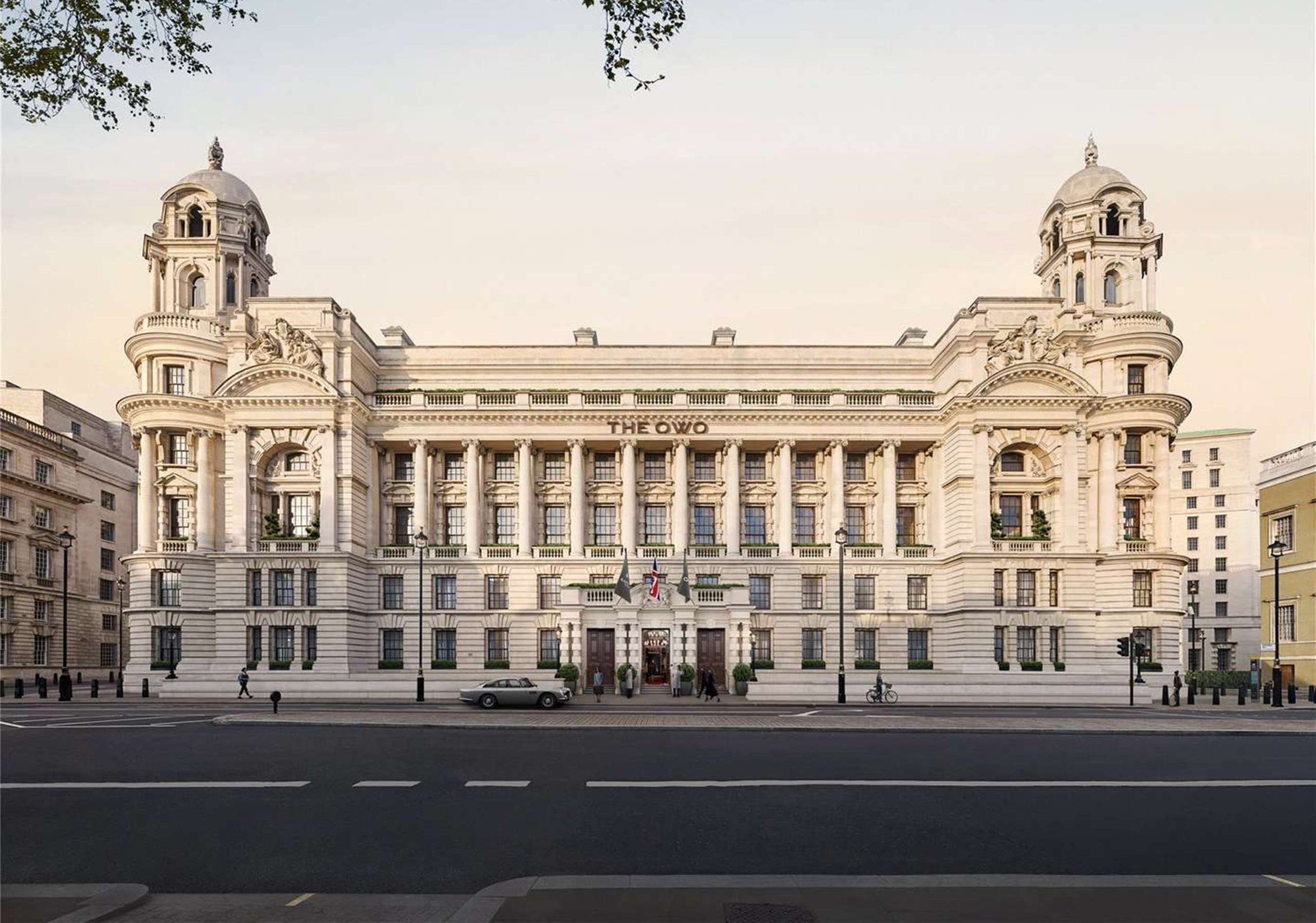 The OWO Residences by Raffles exteriors. Majestic neoclassical building with grand columns, ornate facades, and two towers; the setting is serene with a pale sky and sparse traffic.