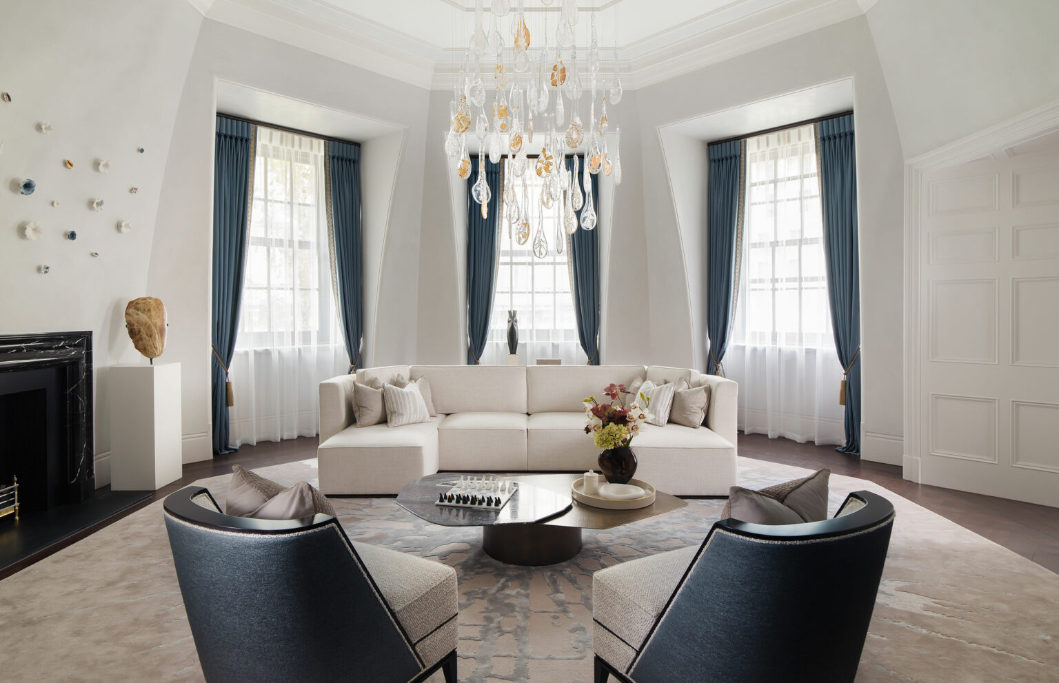 Elegant living room with a white sectional sofa, blue curtains, and a round coffee table displaying a flower arrangement. A modern chandelier adds luxury.