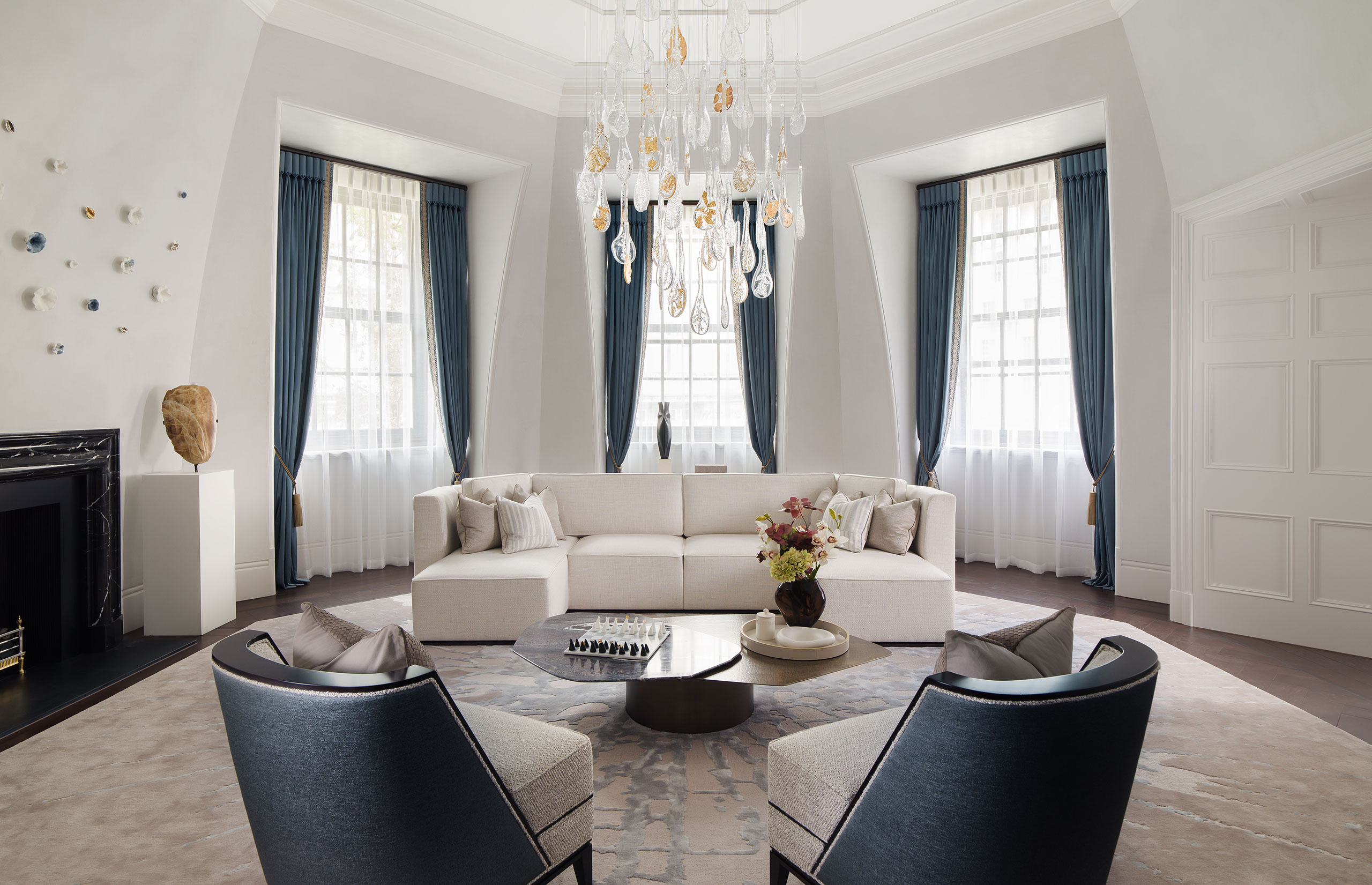 Elegant living room with a white sectional sofa, blue curtains, and a round coffee table displaying a flower arrangement. A modern chandelier adds luxury.