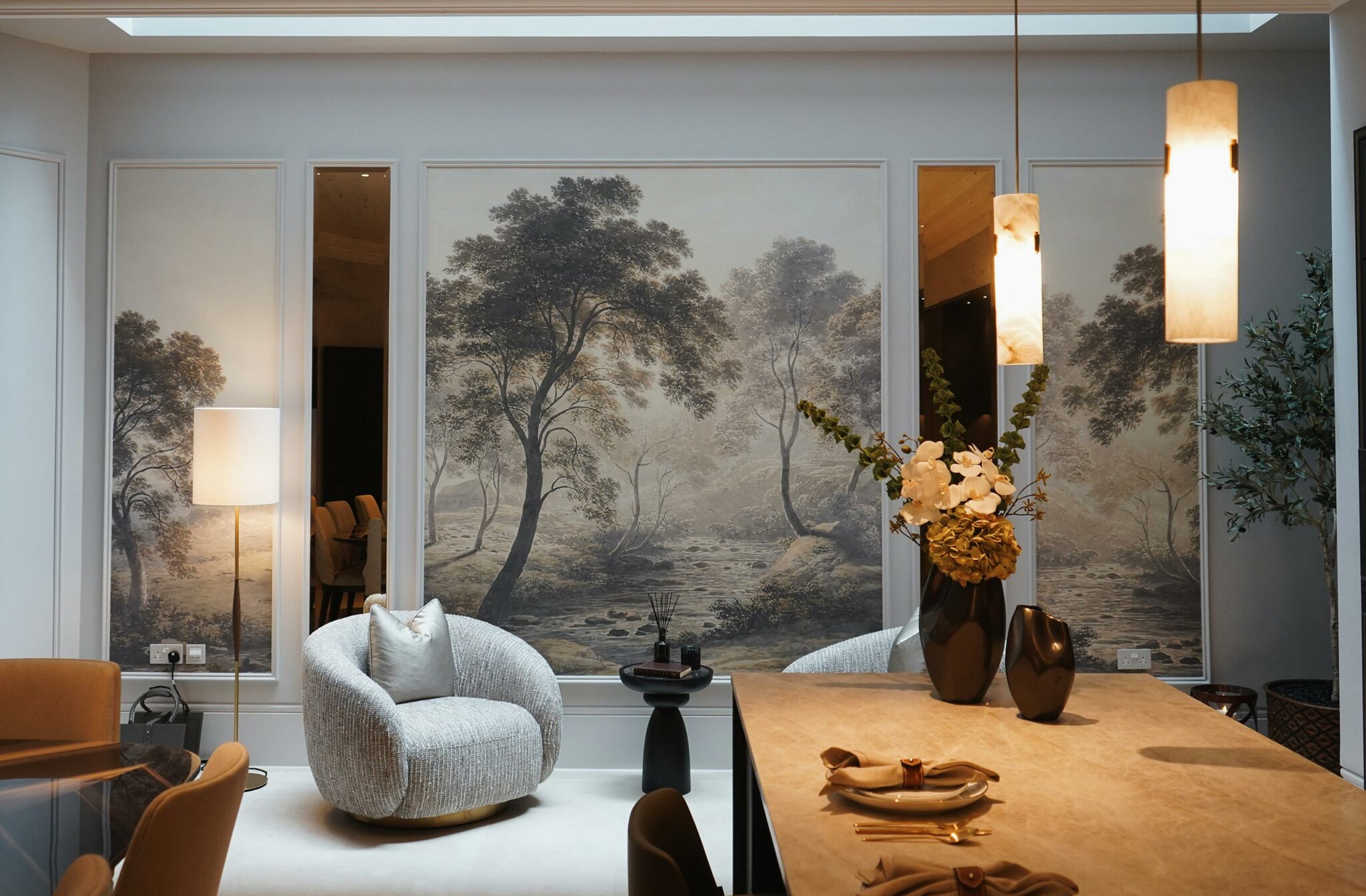 Sophisticated living room with a serene forest mural, plush gray chair, warm lighting, and elegant table setting. Mood is calm and inviting.