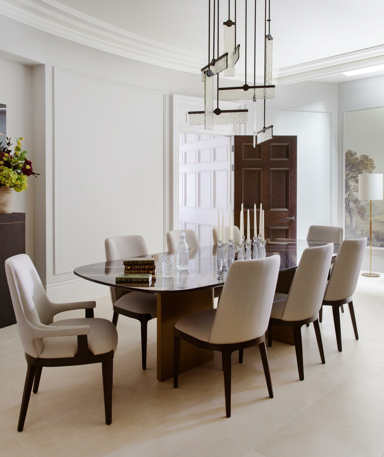 Elegant dining room with a dark oval table, eight cushioned chairs, and candles. Modern chandelier, floral arrangement, light walls, and artistic floor lamp.