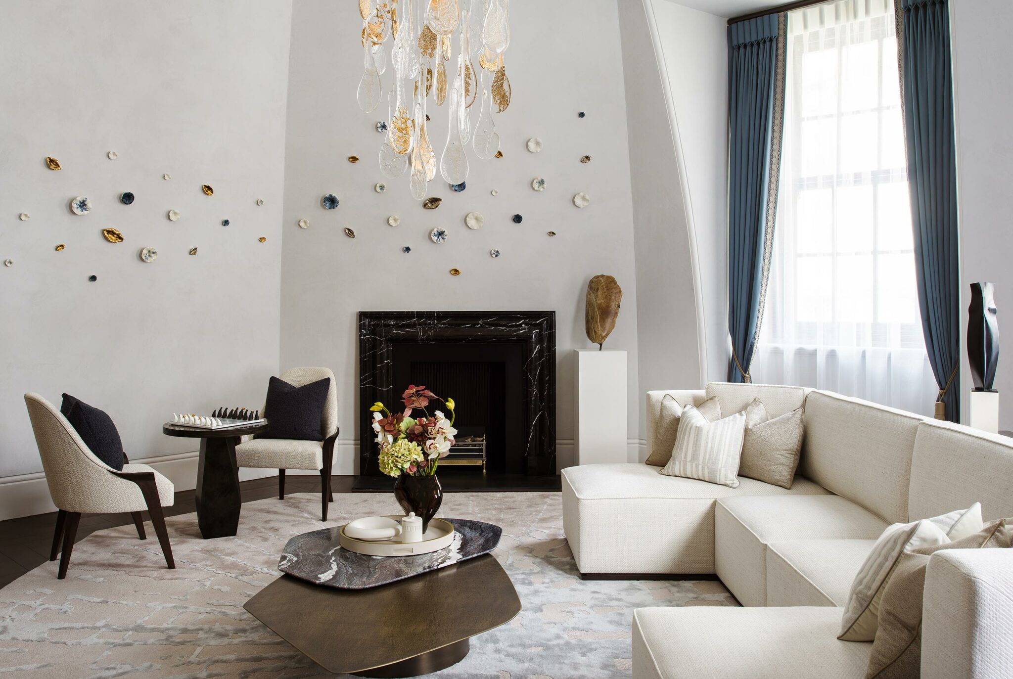Elegant living room with a marble fireplace, abstract wall art, plush white sectional, blue curtains, sculpted chandelier, and floral centerpiece. Cozy and sophisticated ambiance.