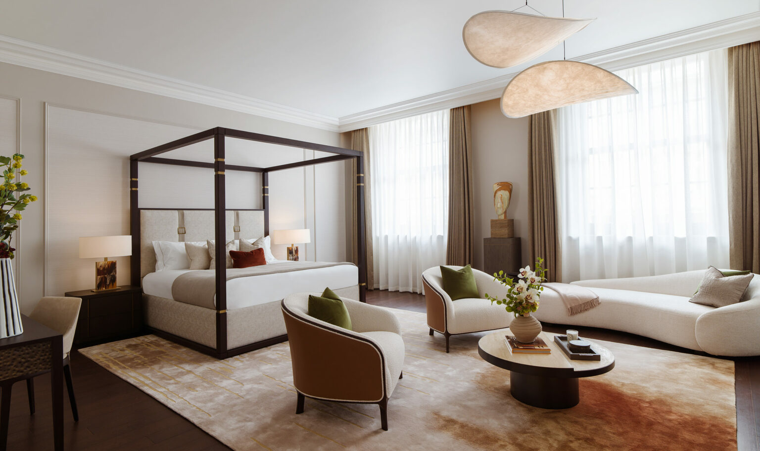 Elegant bedroom featuring a large canopy bed with white linens and brown accent pillows. Two plush armchairs with green cushions face a round coffee table, exuding a cozy, sophisticated atmosphere.