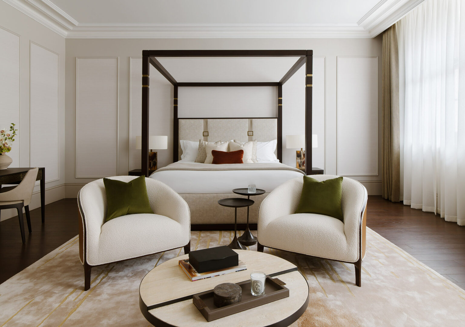 Elegant bedroom featuring a large canopy bed with white linens and brown accent pillows. Two plush armchairs with green cushions face a round coffee table, exuding a cozy, sophisticated atmosphere.