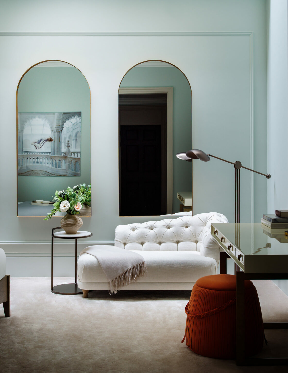 Elegant living room with a white tufted chaise lounge, a vase of flowers on a side table, arched mirrors, soft lighting, and a tranquil, sophisticated ambiance.