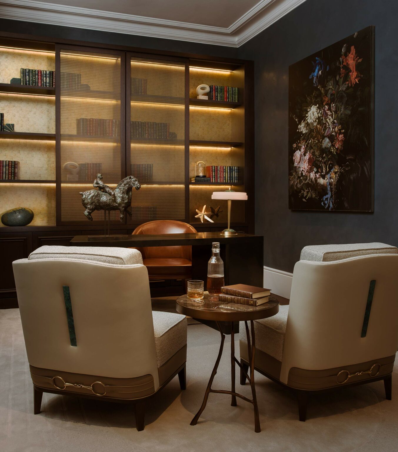 Elegant study with two beige chairs facing a wooden desk. Backlit bookshelves, a horse sculpture, a painting, and warm lighting create a cozy ambiance.