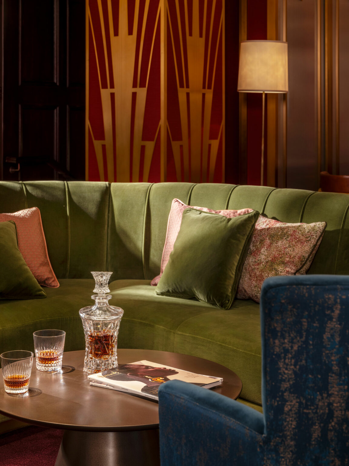 A cozy, elegant private club lounge nook with a green velvet couch adorned with pink and green pillows. A crystal decanter with whiskey and glasses sits on a round table, alongside a magazine. The backdrop features art deco patterns in warm tones.