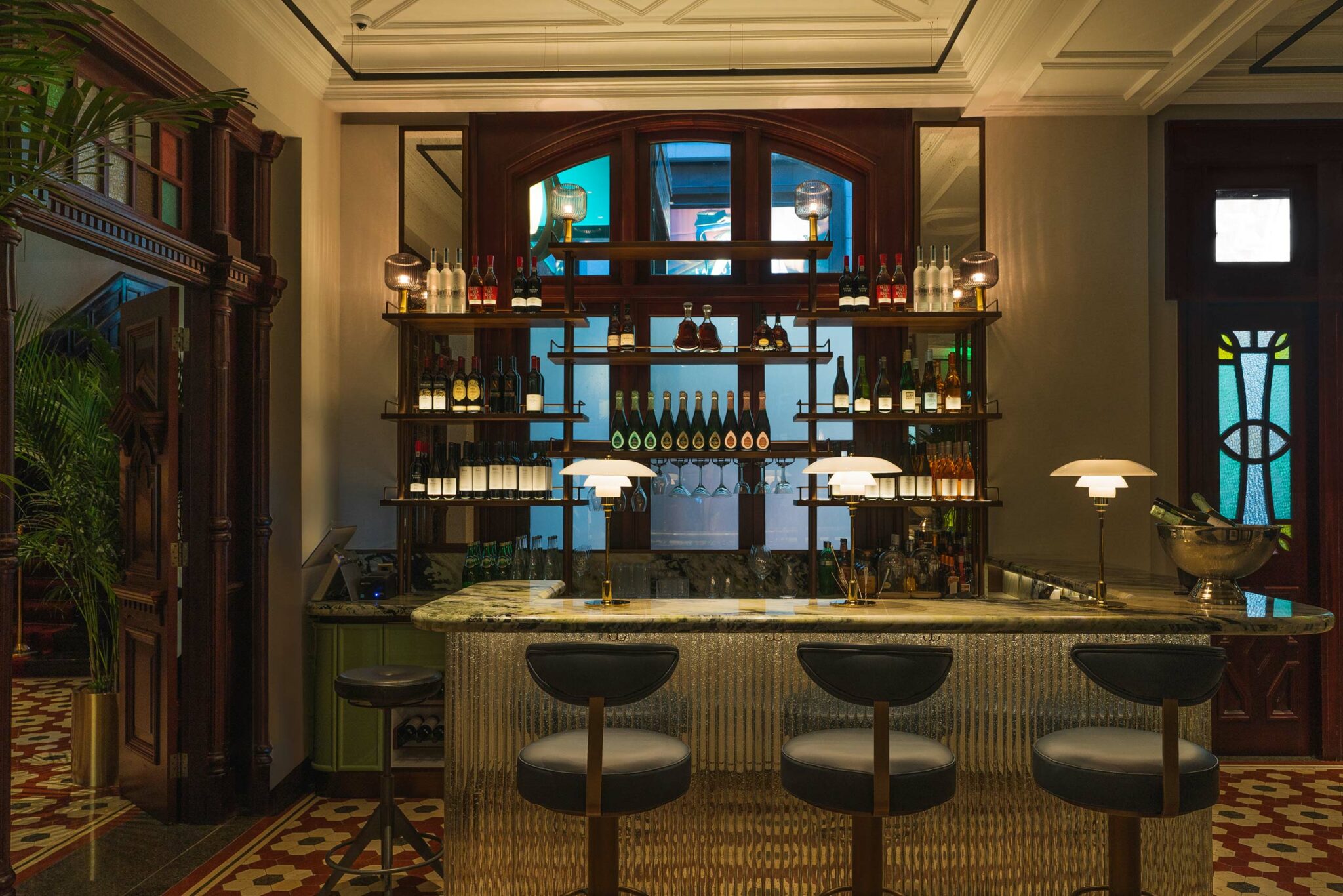 Elegant private club bar with marble countertop, three round chairs, and illuminated shelves filled with wine bottles. Warm lighting creates a cozy ambiance.