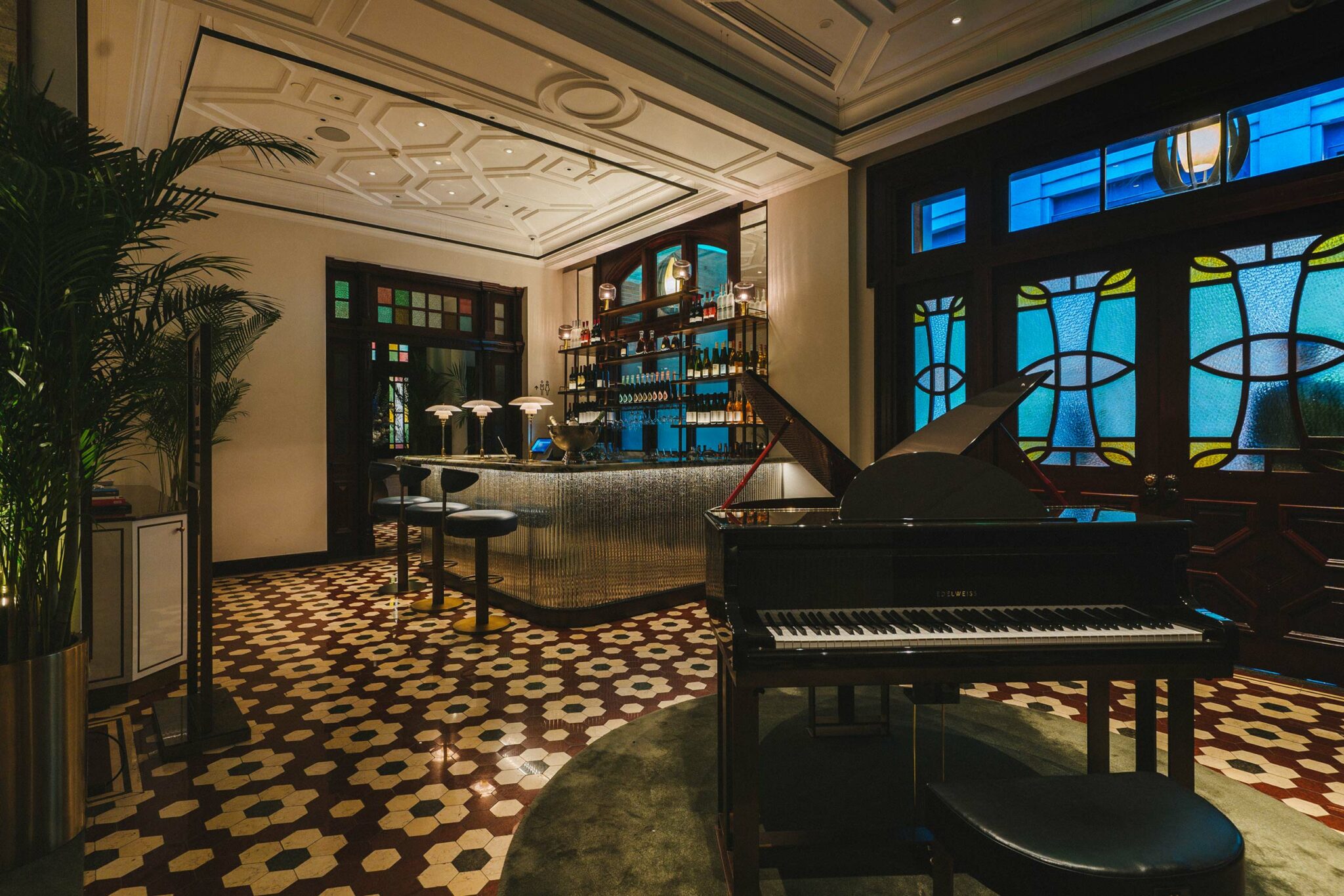 Private club chic bar with a glossy black piano on a geometric tiled floor. Stained glass windows and dim lamps create an elegant, sophisticated ambiance.