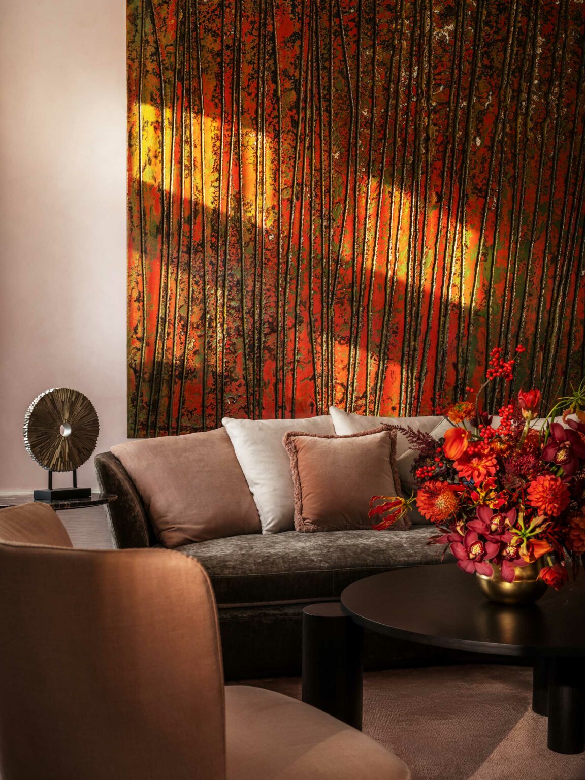 Warm-toned private club private room with an abstract orange, yellow, and green mural, a plush sofa with cushions, a round table with vibrant red flowers, and a decorative sculpture.