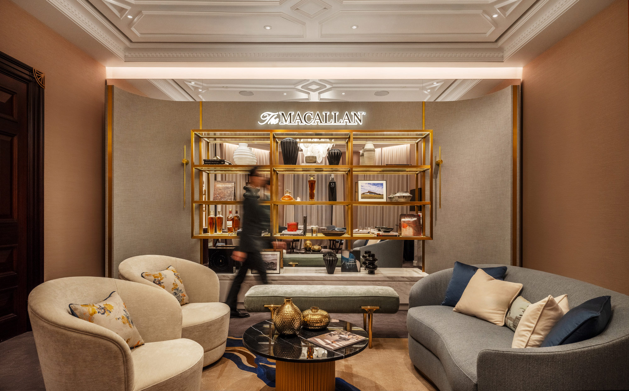 Luxurious private club private room with elegant sofas and decorative pillows. A well-dressed person walks past a display of shelves with bottles and art. Sofisticated ambiance.