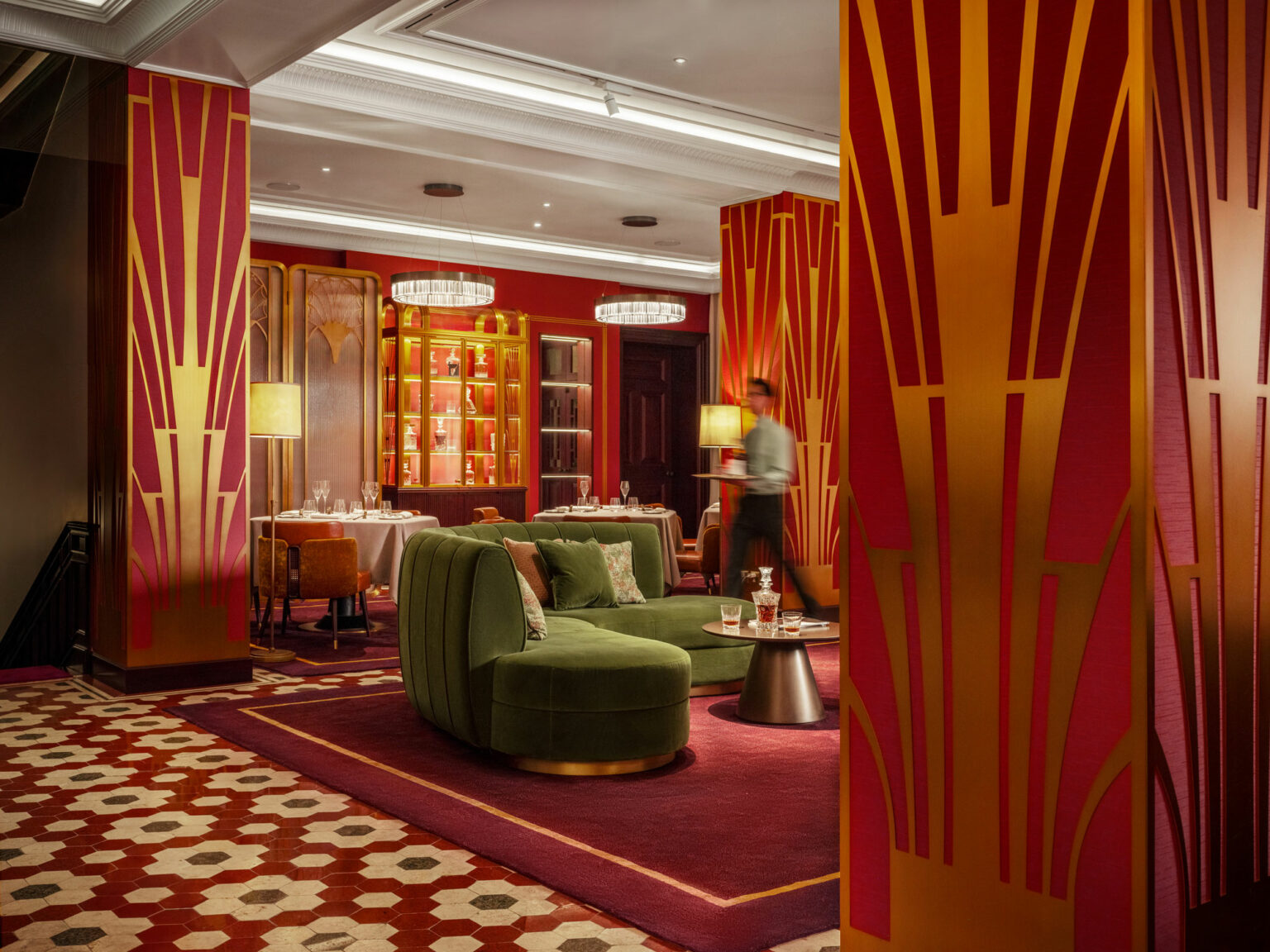 Art deco-inspired private club lounge with rich red and gold walls, green velvet sofas, and geometric patterns. Elegant ambiance with warm lighting and artful decor.