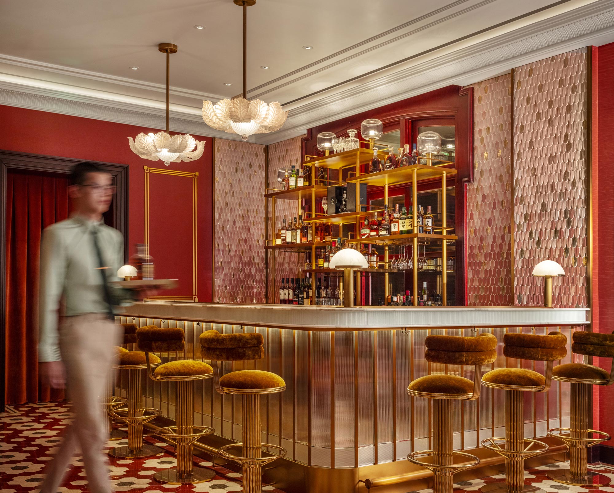 Sophisticated private club bar with velvet stools, gold accents, and intricate lighting. A server in motion enhances the elegant, lively ambiance.