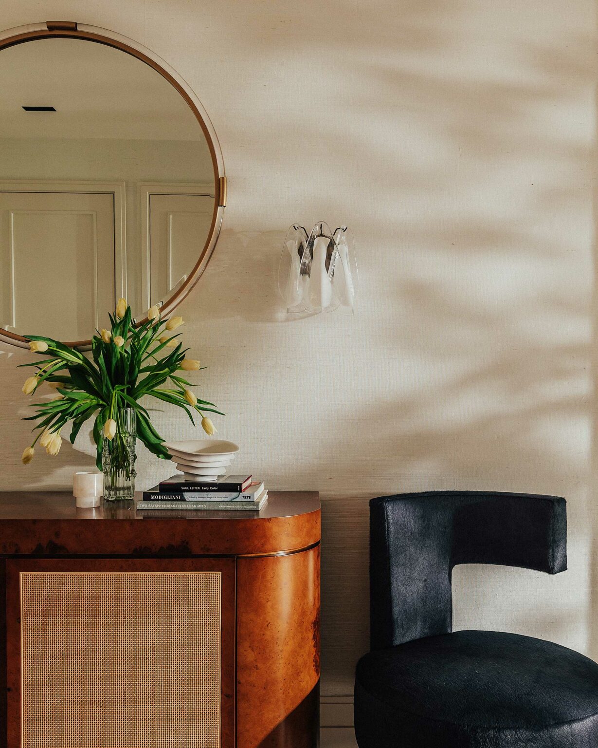 Warm interior with a round mirror, a vase of tulips on a wooden console, and a dark chair. Soft light creates a serene, elegant ambiance.