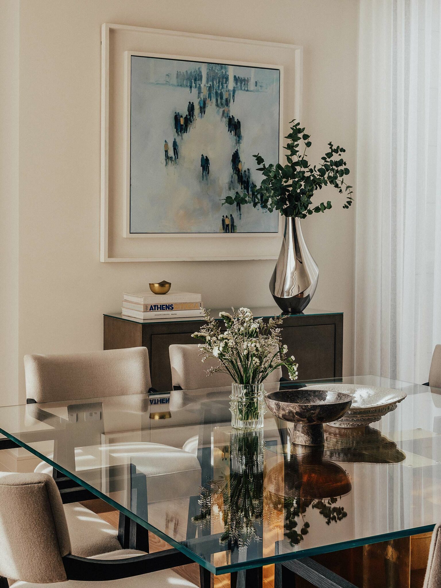 Modern dining room with a glass table, beige chairs, and a vase of wildflowers. A large painting and sleek silver vase add artful elegance.