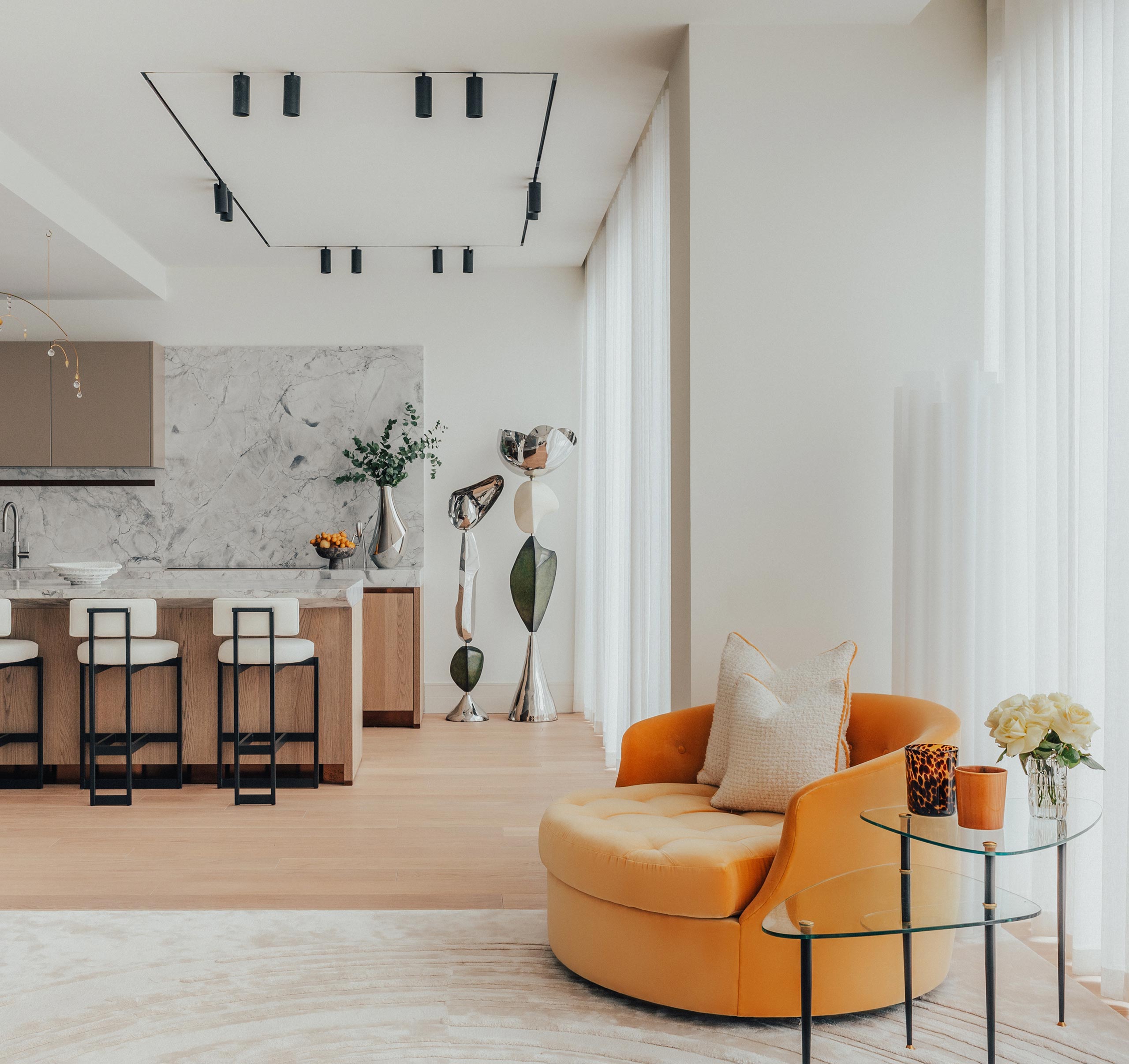A modern living room with a cozy, round orange chair beside a glass table. The open kitchen area features marble accents and bar stools, with a sleek, contemporary sculpture nearby.